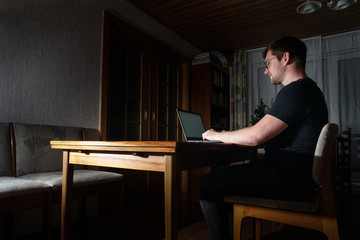 Mann in schwarzem T-Shirt sitzt alleine im dunklen Raum am Tisch aus Holz und arbeitet am Laptop....