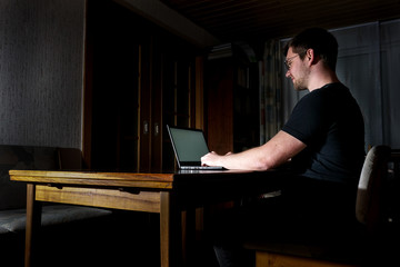 Mann in schwarzem T-Shirt sitzt alleine im dunklen Raum am Tisch aus Holz und arbeitet am Laptop....