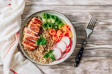 Healthy buddha bowl lunch with grilled chicken, quinoa, cucumber, radish, carrots on white wooden background.