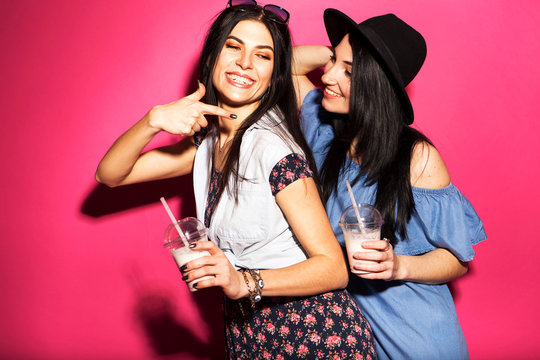 Two caucasian brunette hipster woman in casual stylish outfit having fun drinking milkshakes with straws. They standing on a bright pink background. Cheerful, happy emotions