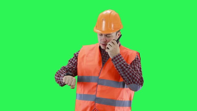 A man in a construction helmet and an orange vest looks at his watch, talking on the walkie-talkie,green screen background