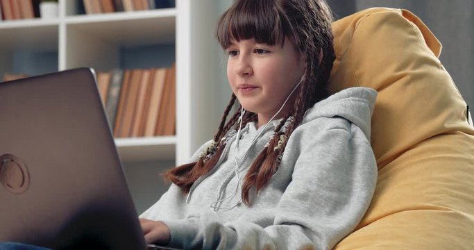 Pretty young girl with trendy hairstyle sitting in modern armchair bag and having video chat on personal laptop. Female teenager in domestic outfit studying remotely at home.