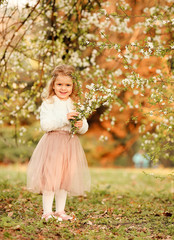 A beautiful cute little girl laughs and has fun in a flying nude skirt and a white fur blouse with a bouquet of blossoming cherry flowers in a spring botanical garden. For Easter and spring cards