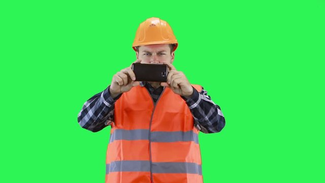 A man in a construction helmet and orange vest shoots video on a smartphone, green screen background