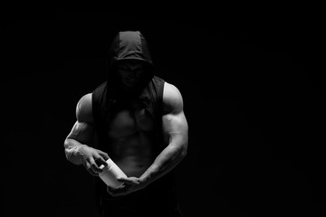 Naklejka na ściany i meble Muscular bodybuilder with jar of protein on a dark background. Sports nutrition. Bodybuilding nutrition supplements, sport, workout, healthy lifestyle concept. Black and white photography