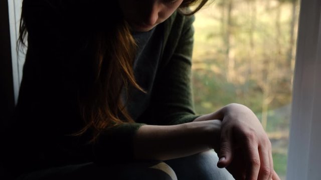 Close up of thoughtful girl sitting on sill with folded hands on knees looking sad at window, depressed woman spending time alone at home, young upset pensive woman feeling lonely. Selective focus