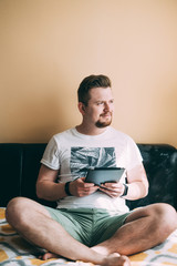 A man with a beard in shorts and a white T-shirt is sitting on the bed with a tablet and looking...