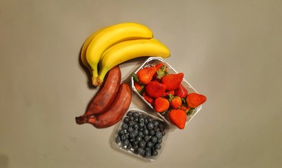 owoce ,na szarym tle ułożone zółte i czerone banany obok pojemnik z czerwonymi dorodnymi...