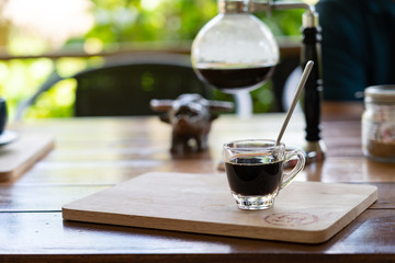 glass of coffee on wood plate with blur coffee maker behide