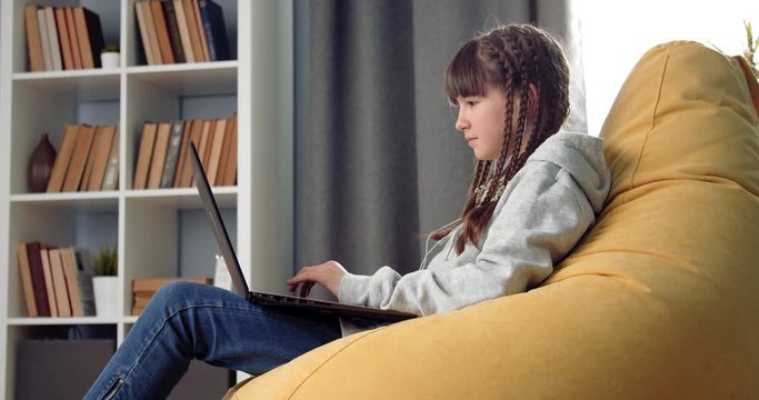 Side view of happy young girl with trendy hairstyle doing homework on laptop while sitting on comfortable beanbag chair. Concept of remote studying and online education.
