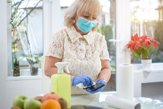 Mid Age Woman Wearing Protective Mask And Gloves Disinfecting Her Phone With Paper Tissue At Home