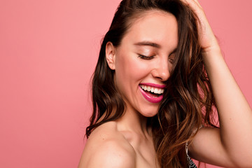 Studio shot of attractive laughing girl with wavy brown hair with nude make up, pink lipstick posing at camera with closed eyes over pink background
