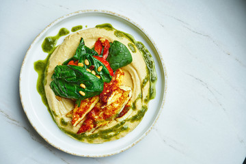 Traditional oriental appetizer - chickpea hummus with fried halumi cheese, spinach, tomatoes and olive oil on a white ceramic plate on a marble background