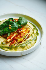 Traditional oriental appetizer - chickpea hummus with fried halumi cheese, spinach, tomatoes and olive oil on a white ceramic plate on a marble background