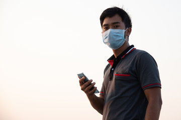 silhouette portrait of Asian man wearing medical mask and dark gray shirt holding phone by right hand and looking straight in evening sky background stock photo