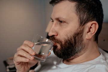 A lonely bearded dark-haired man drinks water from a glass while lying on his bed in the evening. Close-up on the right, healthy habit, bedroom, self-isolation.
