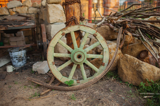 Rustic still life with an old pistachio-colored wooden wheel on a background of branches, stones, an old bucket and garbage in the rays of the setting sun