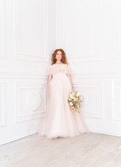 Fototapeta premium Beautiful natural redhead girl bride, with nude makeup, wearing a white dress, holds a wedding bouquet in her hands, standing an the wall in a light interior.