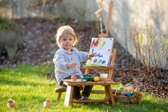Sweet toddler child, painting eggs in garden with little chicks running around him