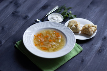 White plate with cabbage vegetarian soup on table cloth, baked buns, sour cream, dill and parsley  on black wooden background