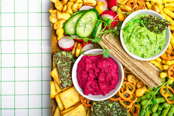 Vegan charcuterie board with beetroot and green pea hummus, various crackers, fresh vegetables. Closeup view, horizontal image