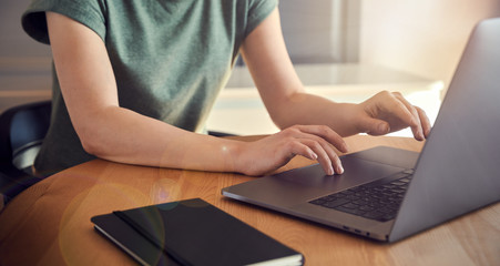 Young woman in green t-shirt sitting at the wooden table at home with laptop and notebook, working...