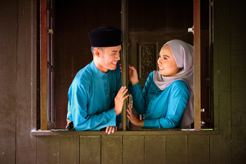 Obraz na płótnie Canvas Young couple of malay muslim in traditional costume having romantic conversation during Aidilfitri celebration at wooden window of traditional house