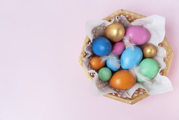 Fototapeta na wymiar Golden, pink, blue, green, orange eggs in the wicker basket on the light pink background. Copy space. Place for text and design. Happy Easter. Top view. Flat layout.