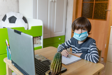 Distance learning for children during the coronavirus epidemic. A boy sits at a table and performs tasks of a teacher on the Internet.