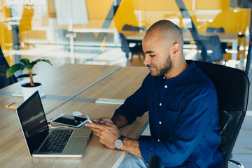 Handsome cheerful african american man in creative atmosphere using laptop sitting on a wooden table.