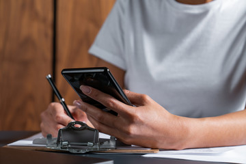 Busy young woman signing papers at office table and double checking information on mobile phone, modern workplace, smart devices.