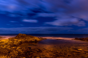 Long exposure shot of a cliff full of stones and a lagoon beside the ocean during a deep night as water poured onto the stones and the surrounding area.