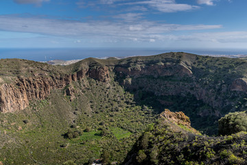 View of the hilltop on a mountainous landscape of volcanic origin along with lots of trees on the horizon, one can see the setting moon of the Gran Canary island