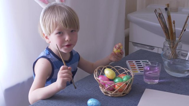 Little blond kid boy coloring eggs for Easter holiday in bath room, indoors. Child holding basket with painted eggs. Child having fun and celebrating feast with easter toy bynny