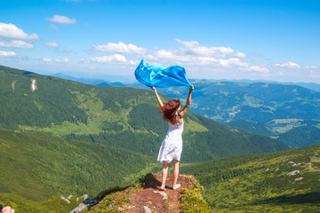 Beautiful young woman in white summer dress stands on a rock and looking into the amazing valley, blue scarf waving on the wind in her hands