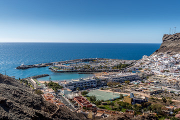 Fototapeta na wymiar View from the air on the coast and bay in the city of Mogan on the Canary Islands where the bay harbor and people walking along the waterfront inside city