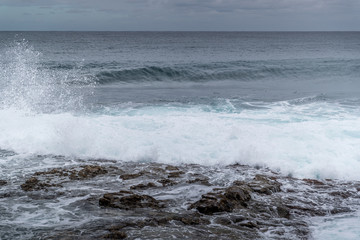 Waves crashing into a rocky shore full of large rocks and breaking water leaping to the sides with a pair of boats moored on the horizon at sea