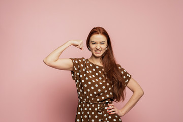 A redhead girl is on the pink background. A redhead girl with the biceps. A strong girl with beautifull red hair on the pink background. A girl is happy with her muscles