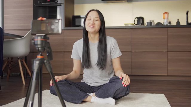 Female Asian lifestyle blogger sitting cross-legged on floor at home, waving at camera and speaking during livestream on cell phone placed on tripod