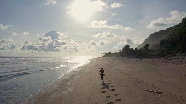 Aerial view sportsman runs alone on sandy beach, ocean and sunset on background