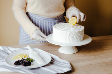Housewife in the kitchen decorates a lemon cake with fruits, blackberry, lemon, mint during quarantine. Recipe of organic homemade lemon cake. Concept of cooking at home