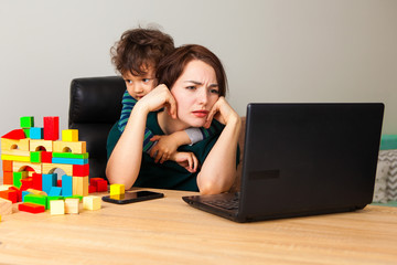 Tired woman at a laptop working at home. A boy, a child assembled a house of cubes and hangs on his...