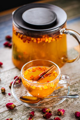 Closeup on the healthy herbal yellow apple tea with sea ​​buckthorn and cinnamon in a glass teapot and mug on the wooden background decorated with roses, vertical