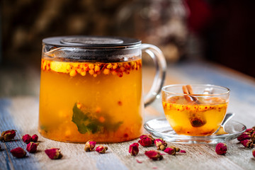Closeup on the healthy herbal yellow apple tea with sea ​​buckthorn and cinnamon in a glass teapot and mug on the wooden background decorated with roses, horizontal