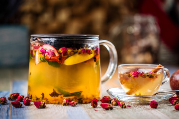 Closeup on the healthy herbal yellow apple tea with passion fruit and cinnamon in a glass teapot and mug on the wooden background decorated with roses, horizontal