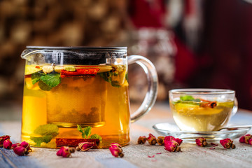 Closeup on the healthy herbal yellow apple tea with mint and cinnamon in a glass teapot and mug on the wooden background decorated with roses, horizontal