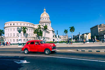 Fototapeta na wymiar Brightly colored classic American cars serving as taxis pass on the main street in front of the Capitolio building in Central Havana, Cuba.