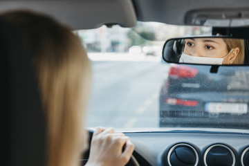 Young woman wearing protective face mask while driving car. Reflection in rearview mirror.  Protection against the novel coronavirus infection.