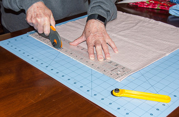 Older woman cutting fabric with a mat board and rotary cutter and clear ruler on the material for sewing or quilting.