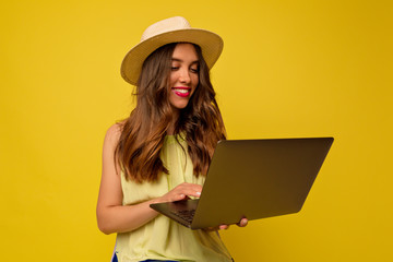 Lovable attractive european young woman with wavy hair wearing hat and summer bright dress working with laptop isolated over yellow background